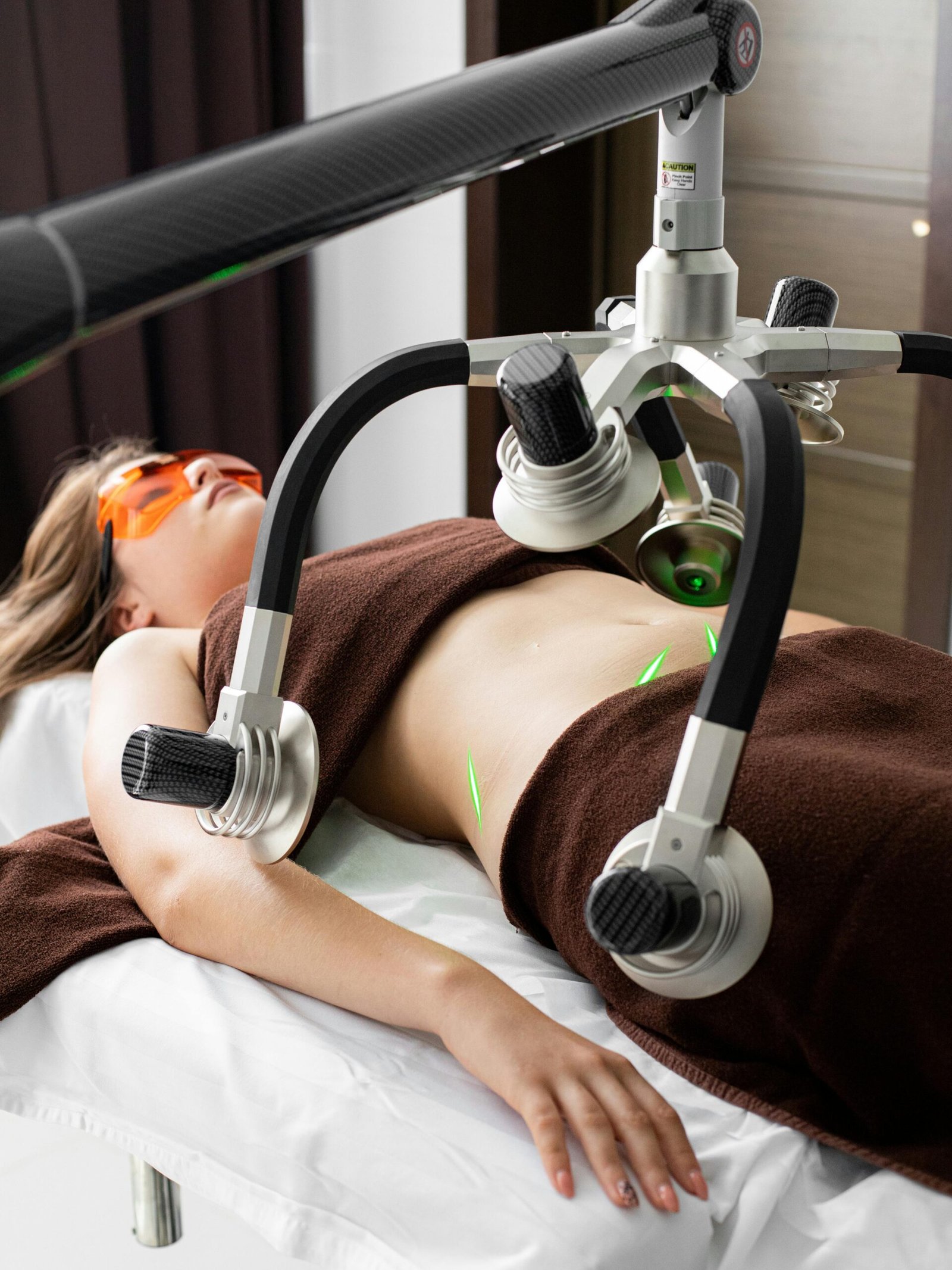 Female client in eyewear lying on table under modern laser fat loss machine during liposuction treatment in light medical clinic